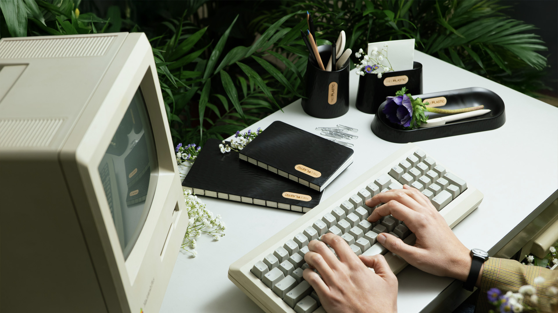 Una scrivania immersa nel verde con un computer vintage e delle mani di donna sulla tastiera. Intorno oggetti di cancelleria in Treeplastic nera di Arbos