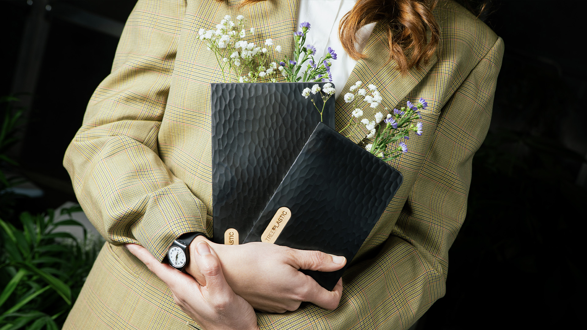 Una donna con un blazer verde chiaro, sorregge due quaderni neri in Treeplastic di Arbos con all'interno dei fiori bianchi