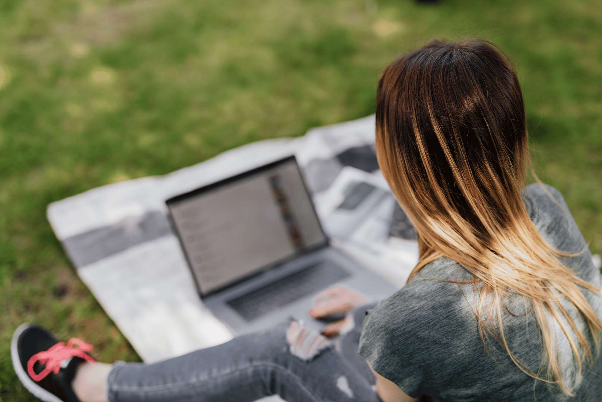 Una ragazza bionda, con jeans, sneakers e maglietta, vista di spalle lavora al pc portatile su un prato